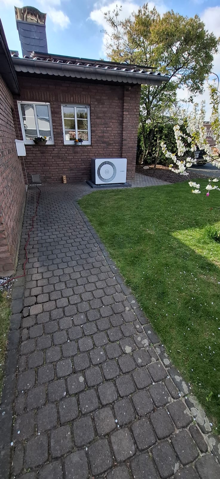 Bosch-Wärmepumpen-Außeneinheit auf Kiesfläche an Backsteinwand unter Fenster, mit sauber verlegten Rohrleitungen.